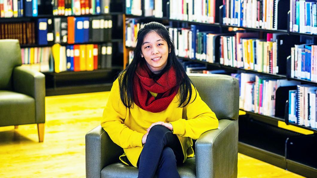 Youth leader Lexin sits in a chair surrounded by shelves of books. She is wearing a yellow sweater and a red scarf.