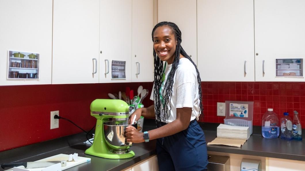 Woman with long braids smiles a she uses a light green mixer in a kitchen