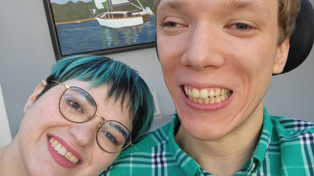 Woman with glasses and turquoise tinged hair smiles with head on shoulder of partner