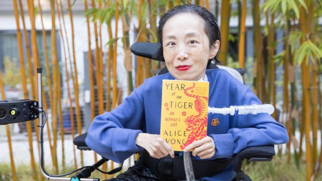 Woman with red lipstick and blue shirt holds orange book in wheelchair