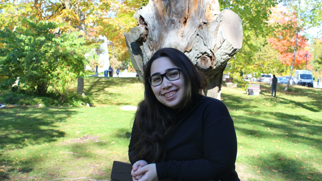 A young person with light skin tone and dark long hair. She is sitting outside in front of a tree and green grass.