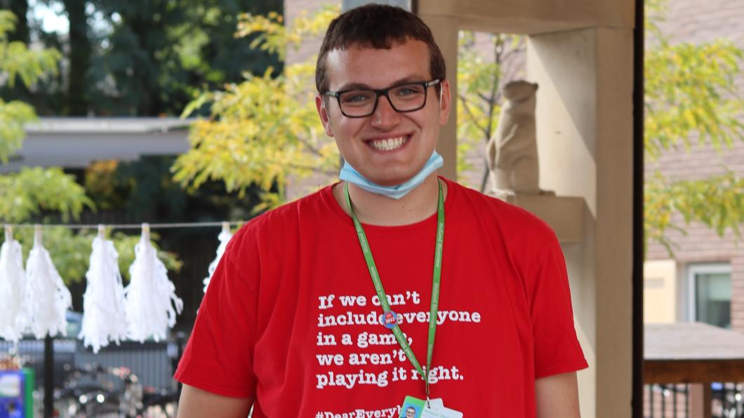 A young person with light skin tone, short brown hair and glasses. They are smiling and standing outside.