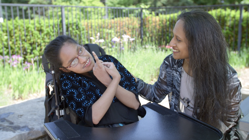 A young girl with medium skin tone and dark hair. She is smiling and sitting next to her mom outside. The young girl is using a wheelchair.