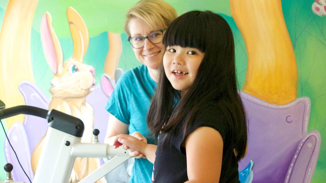 Girl with long black hair with bangs smiles on treadmill with woman with blonde hair holding her hand