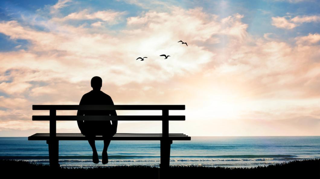 Silhouette of man alone on bench looking out to sea and clouds and seagulls.