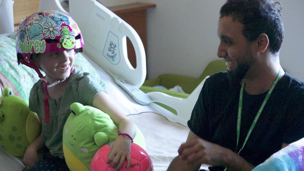 Girl with brightly coloured helmet and holding stuffies smiles on hospital bed at man in black shirt and beard who is also smiling