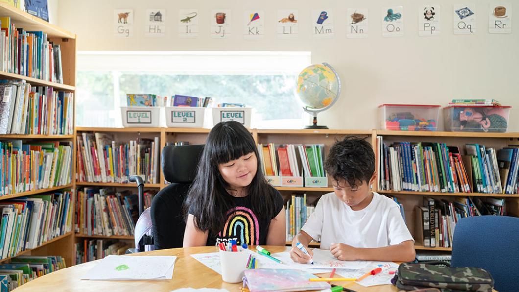 A child using wheelchair on the left, with another child on the right, reading books in a library setting