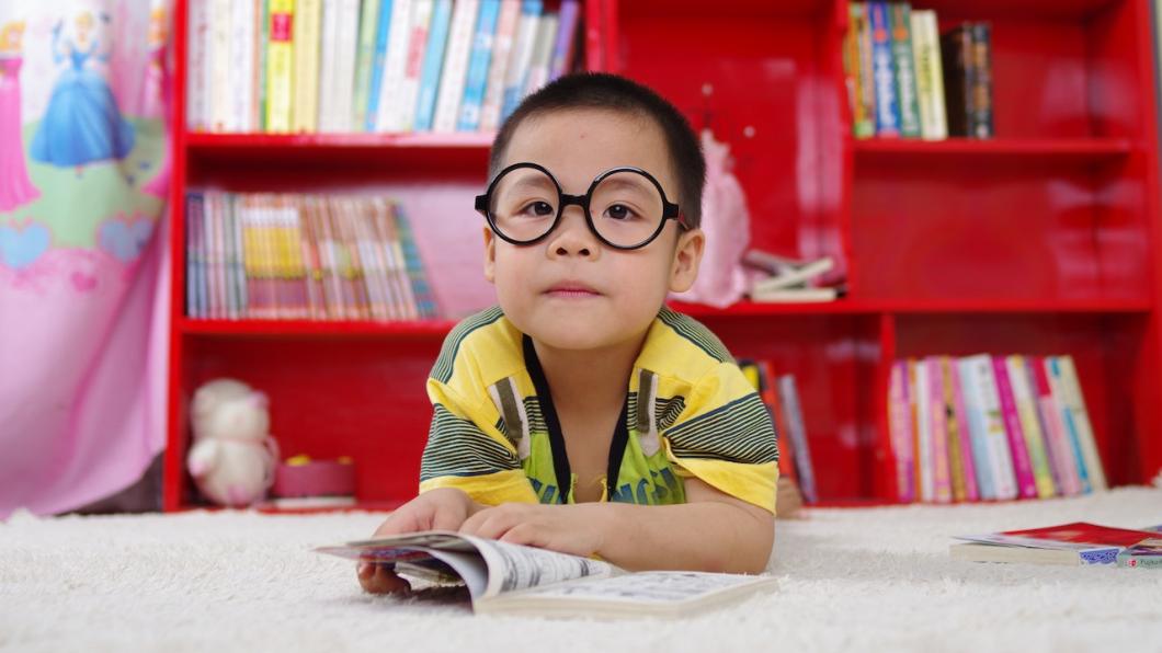 Boy in yellow shirt with big black reading glasses rests on arms on floor with red bookcase behind.