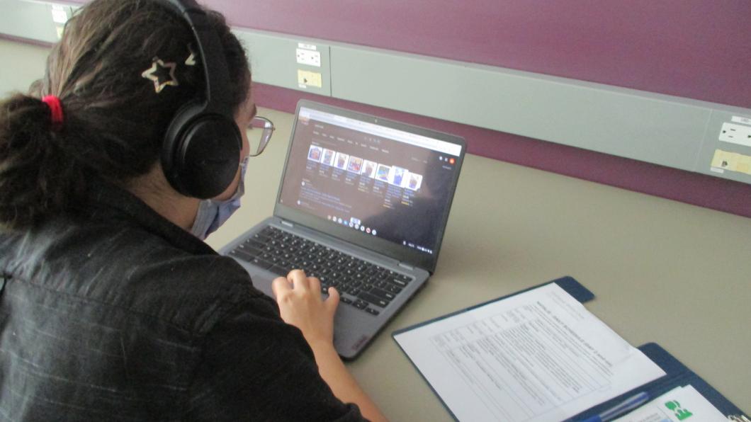 Student with headphones working on a laptop with an open notebook.