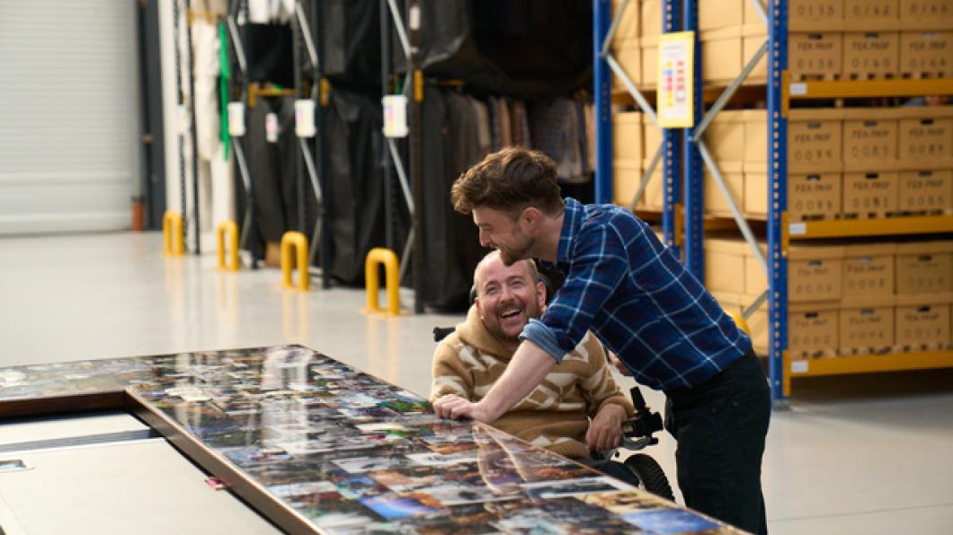 Man stands beside another man in a wheelchair as they look at photos on a large table
