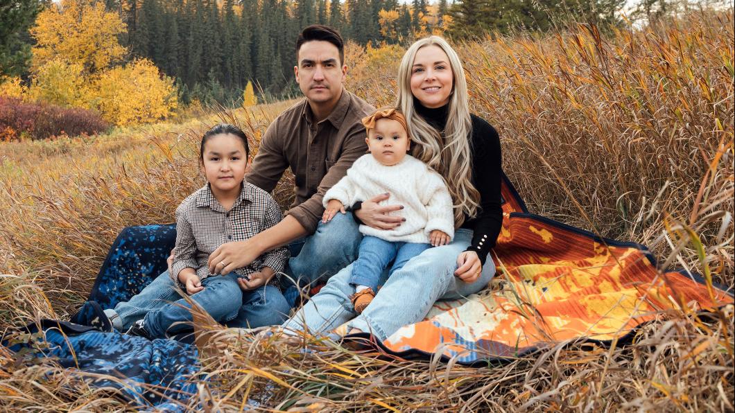 Man with dark hair beside woman with long blonde hair with boy and baby girl in front on a blanket in a field