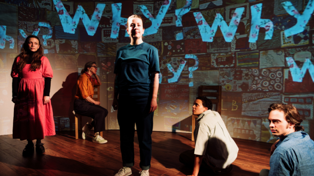 Actors standing and sitting on a stage with the word 'Why?' superimposed in blue on a screen behind them