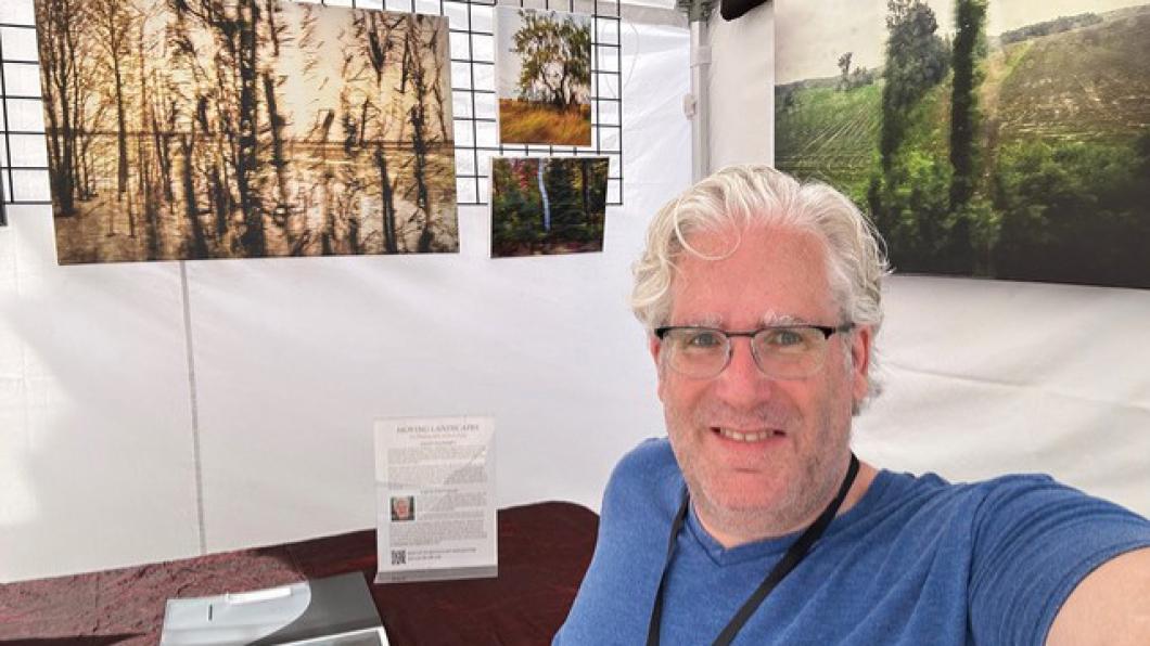 Man with white hair and glasses smiles while sitting in front of an art exhibit