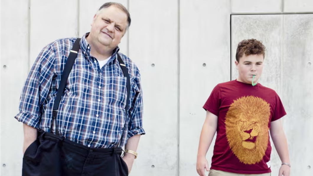 Man in plaid shirt with suspenders stands beside teen with red shirt with gold lion on it
