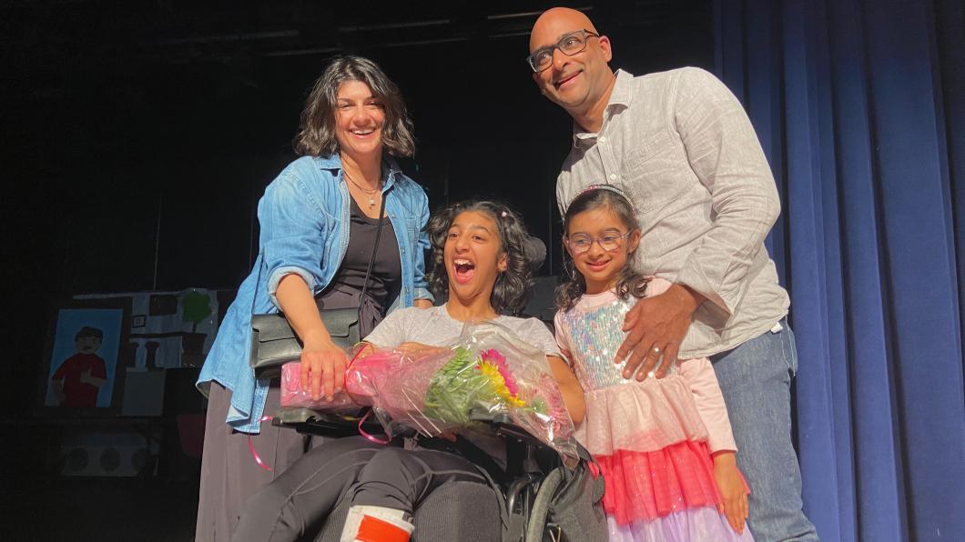 Teen in wheelchair holding flowers on stage surrounded by her family