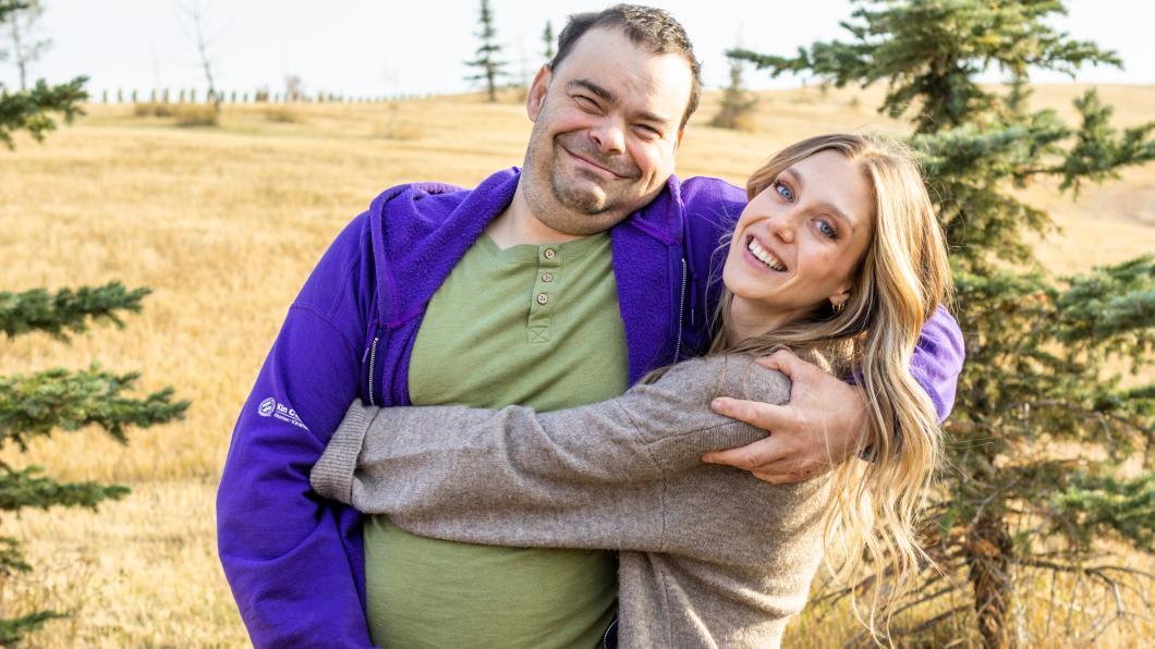 Woman with long light brown hair wraps arms around man wearing a green t-shirt and purple sweatshirt