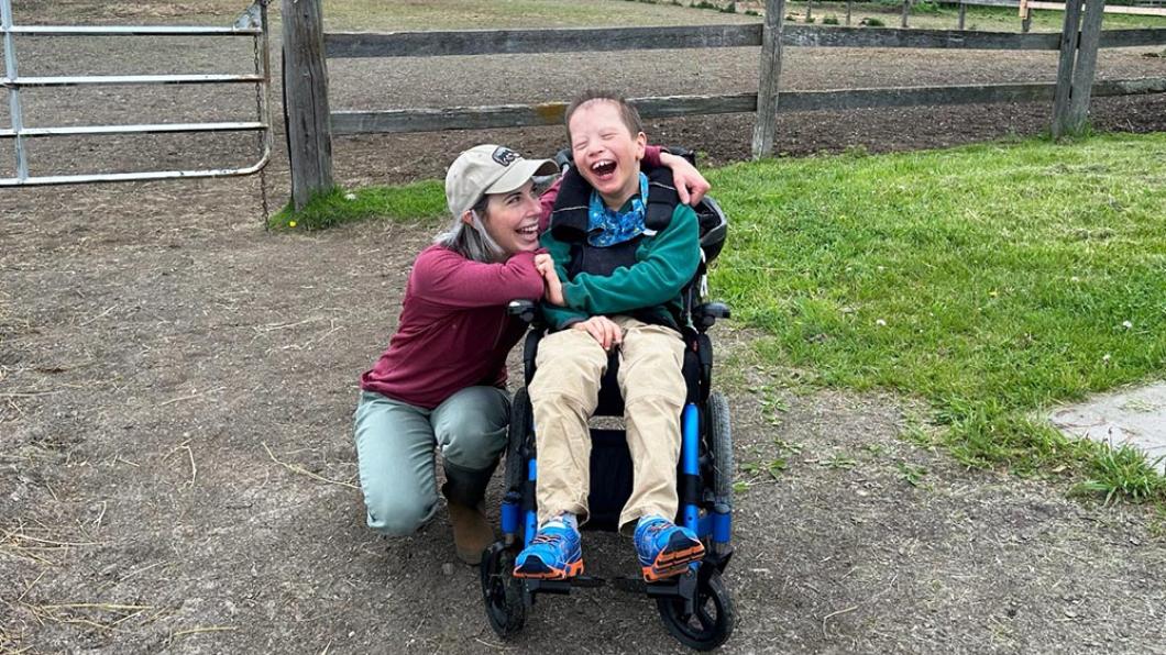 Woman with blonde hair and baseball cap crouches down beside boy in green sweatshirt in wheelchair both smiling