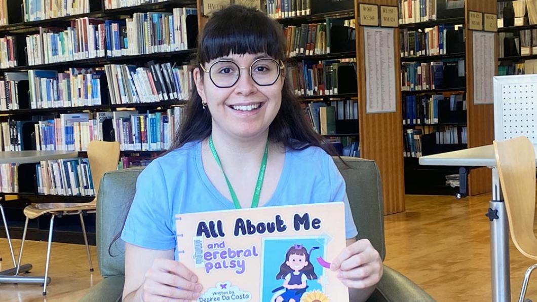 Woman with long dark hair in blue t shirt holds children's book and smiles
