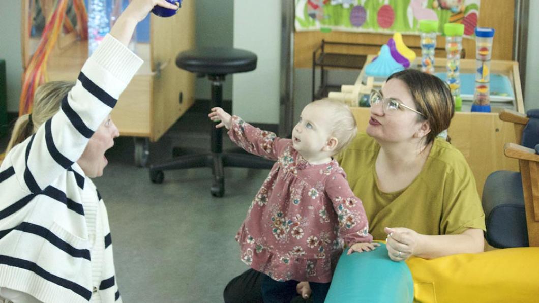 Toddler in pink dress reaches up to a ball held by a woman in a striped sweater while another woman smiles