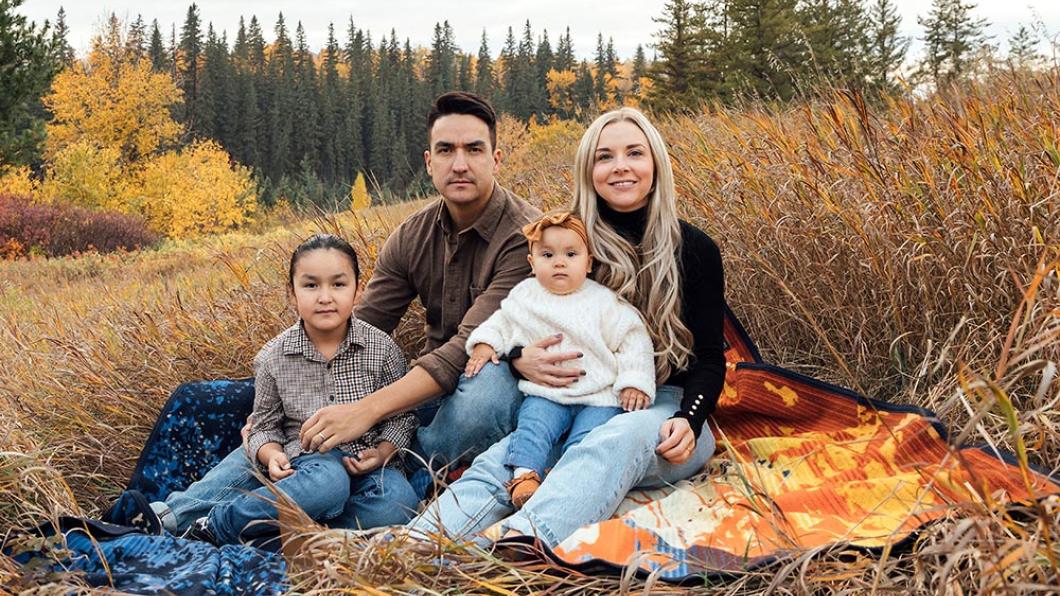 Man with dark hair beside woman with long blonde hair with boy and baby girl in front on a blanket in a field