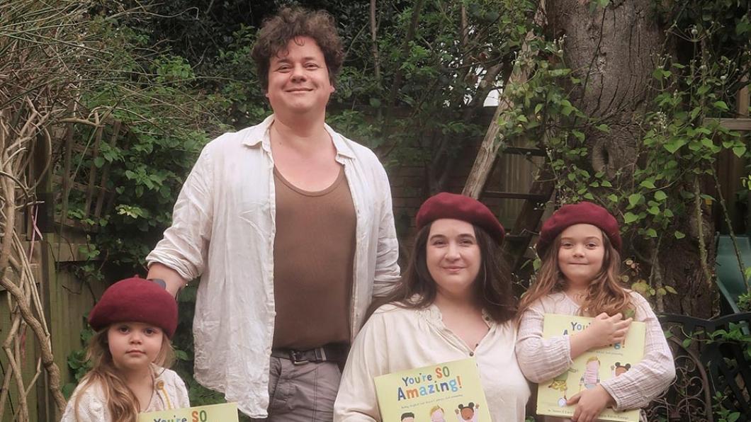A man with brown hair stands behind a woman in a wheelchair and two girls who are each holding a children's book