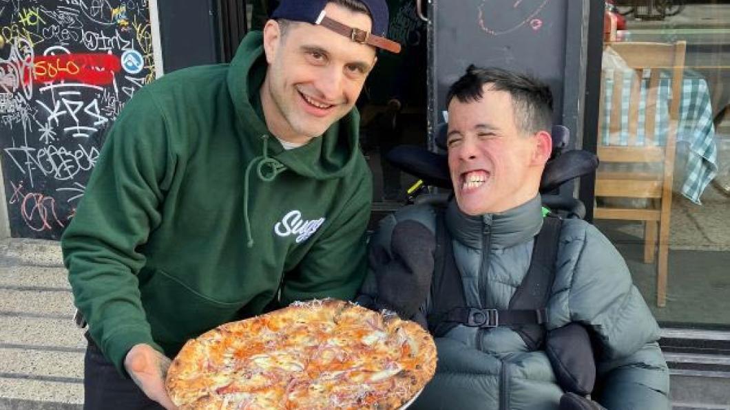 Young man with dark hair grins in wheelchair beside man with basketball cap holding a pizza