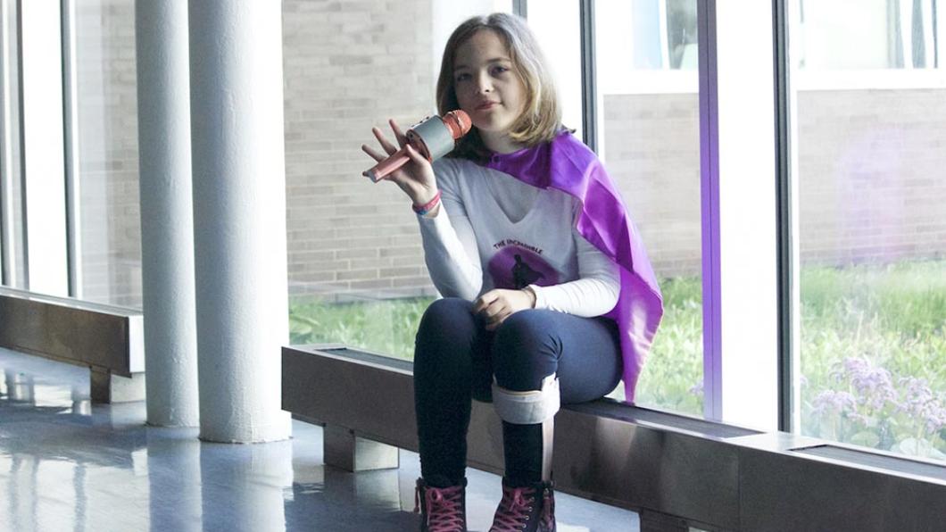 Girl with shoulder length brown hair and pink microphone wears purple cape and sits on bench