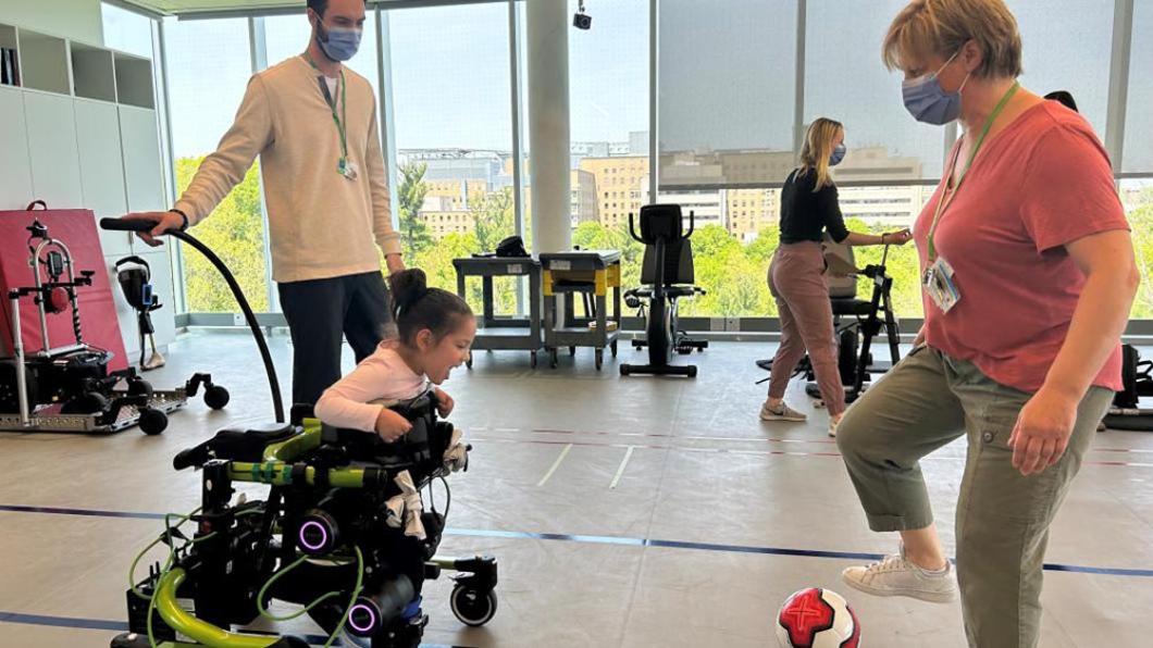 Small girl in green robot-powered walker faces therapist who is kicking a soccer ball