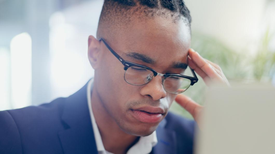 Man wearing glasses with hair in braids and suit jacket looks down with head in one hand