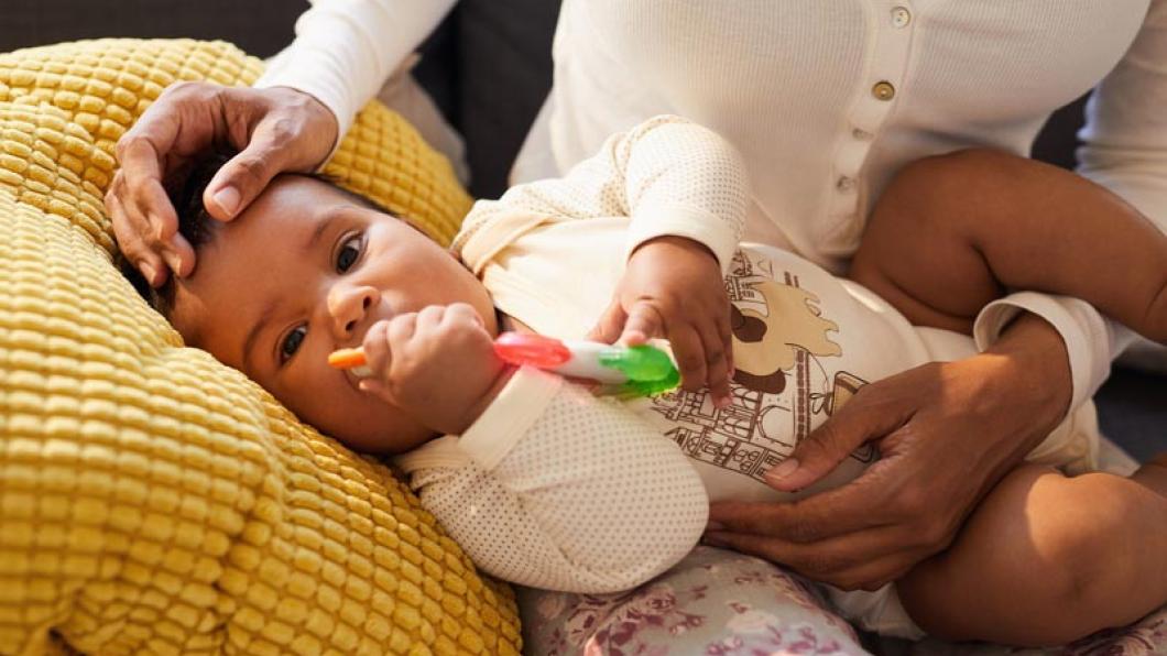 A baby holds an orange and green toy on a pillow balanced on mother's lap