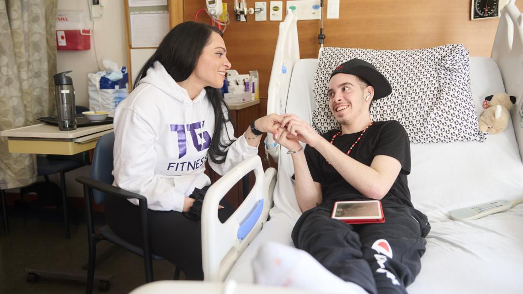 An young person sitting on a hospital bed holds hands with an adult.