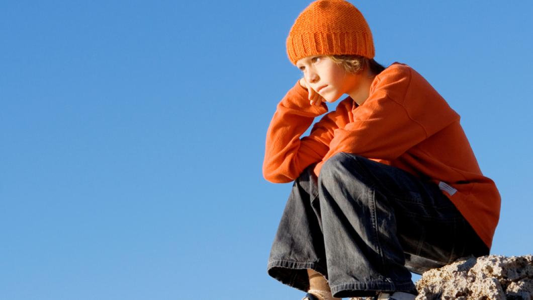 Child in orange sweater and hat sits on rock with head on fist