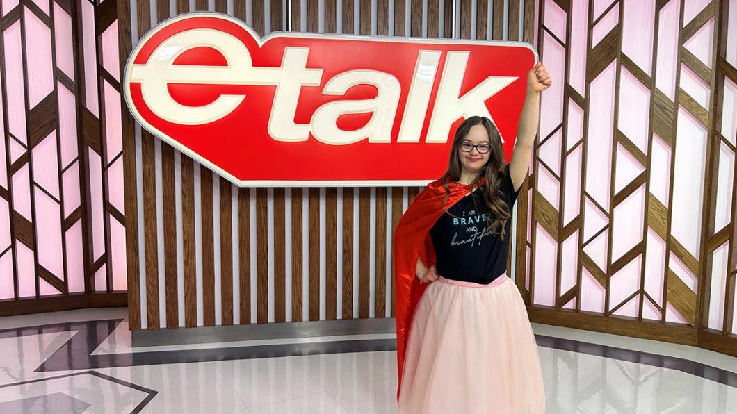 A teenager with a red cape on standing in front of a big "e-talk" logo