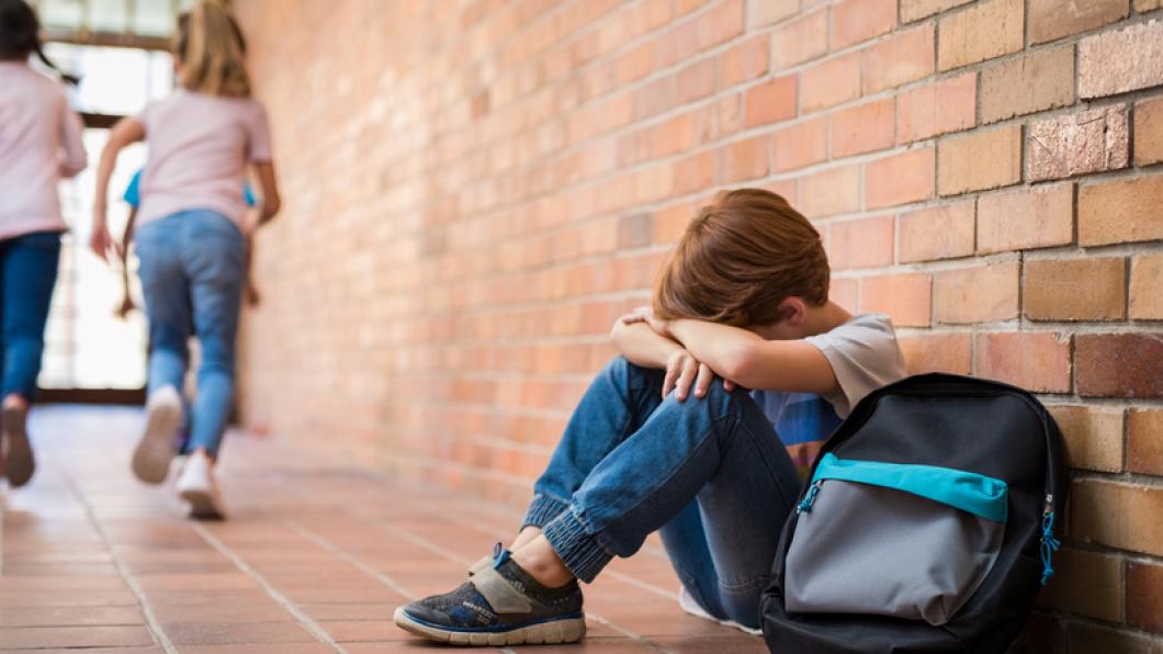 Young boy with head in hands in school hall as other kids run away