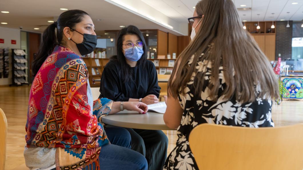 Three adults sit at a table together and have a conversation in the library.