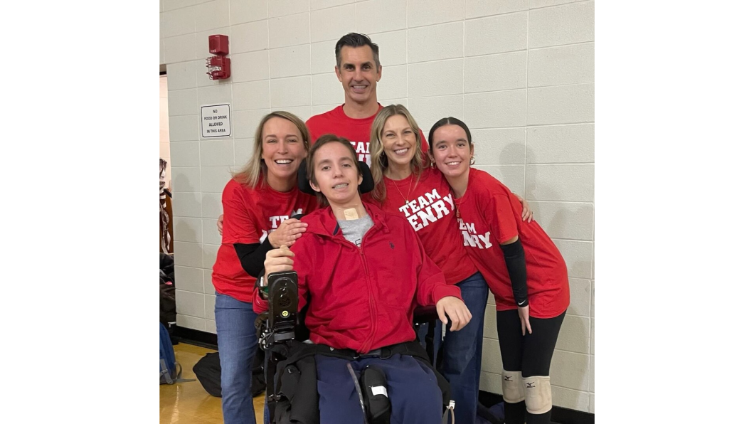 a group of family members huddled around a boy in a wheelchair device. All are wearing red shirts and dark pants. A white background behind them. Everyone is smiling at the camera.