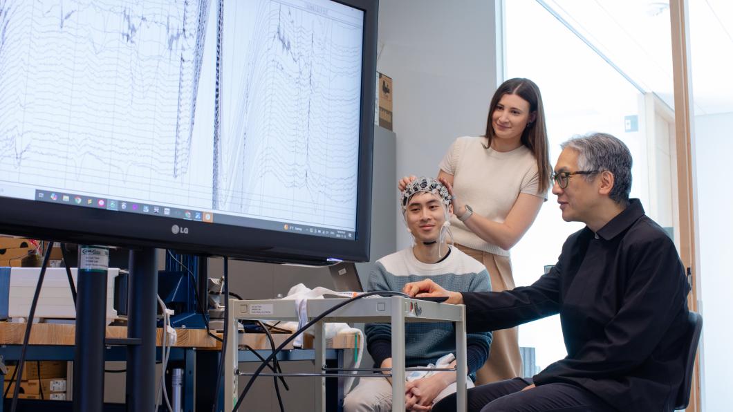 An adult sits and wears a brain computer interface headset while two other adults look at brain wave imaging.