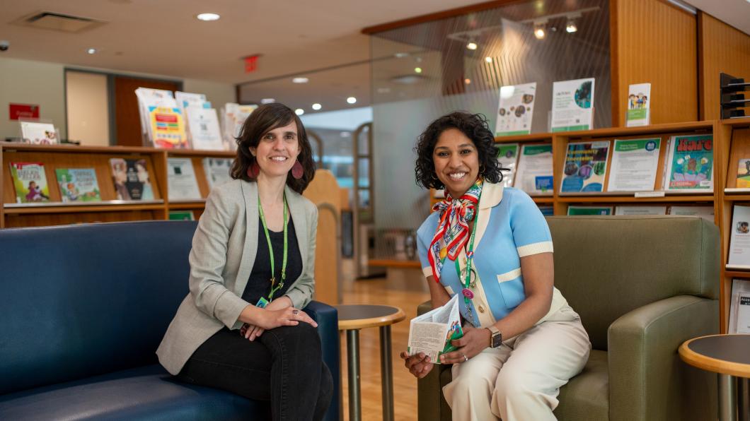 Two adults sit on arm chairs in the library, surrounded by shelves of books and pamphlets.