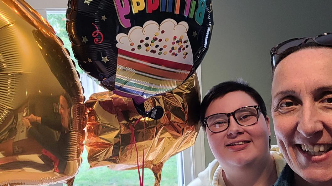 Two women's faces smile beside brightly coloured birthday balloons