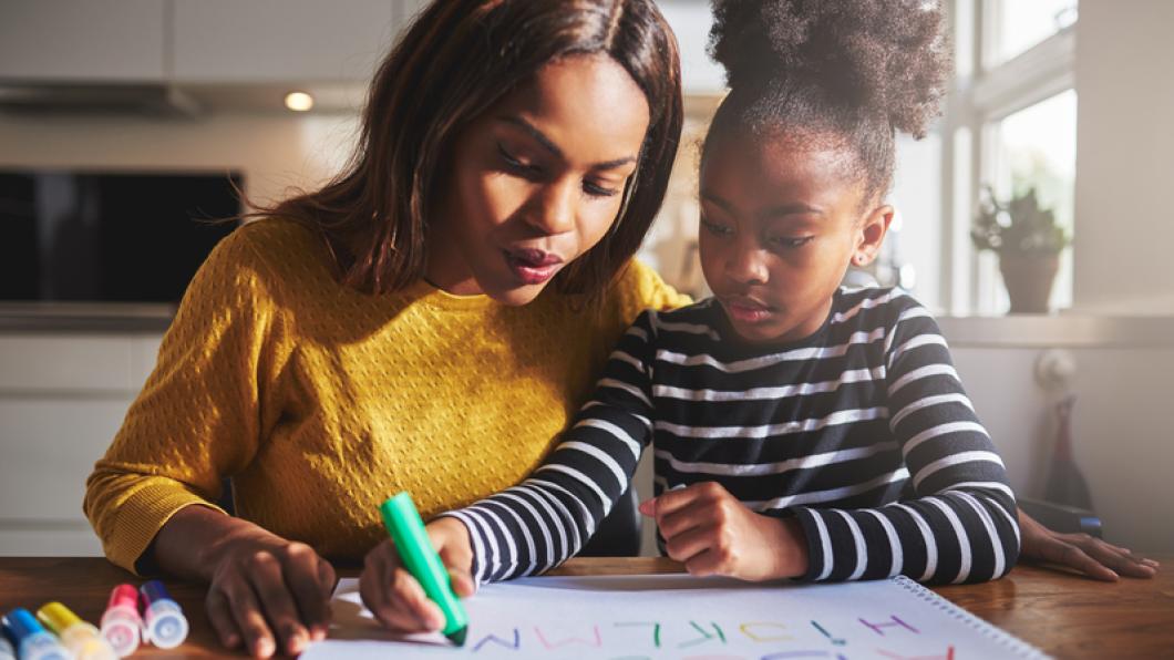 Woman in yellow sweater helps daughter in striped sweater write out alphabet