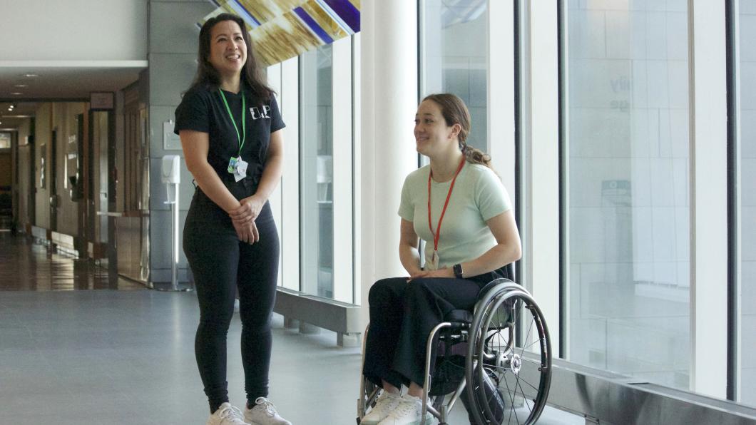 Two women smile beside floor to ceiling window, one in a wheelchair