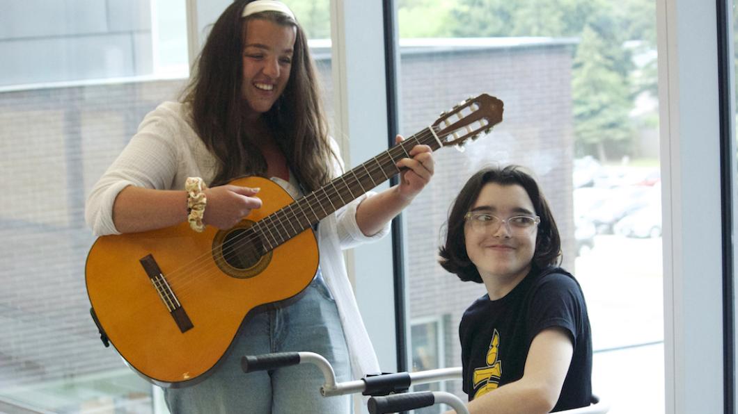 Woman stands playing guitar beside girl sitting on walker smiling