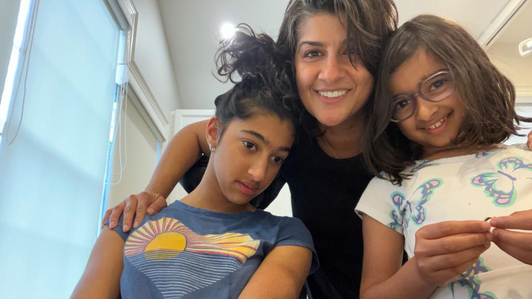 Woman in black shirt smiles with arms around teenage girl in wheelchair and young girl standing