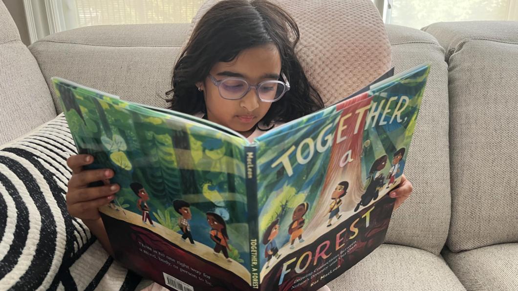 Girl with long dark hair and glasses reads large children's picture book