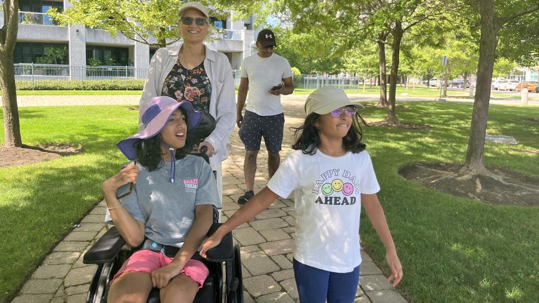 Girl holds hand of teen in wheelchair on path in the park with two people behind them