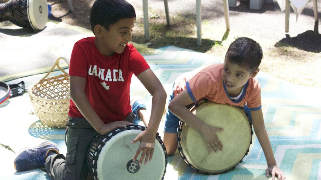 Boy with dark hair and red Canada shirt and boy with dark hair and orange shirt play drums