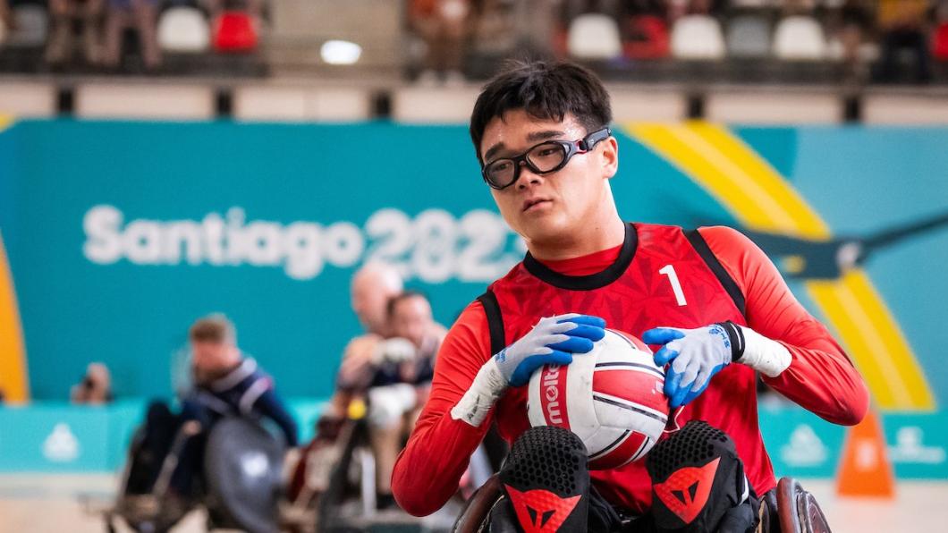 Man with dark hair and glasses wearing red athletic top and gloves holds ball in wheelchair