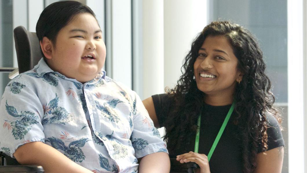Boy with dark hair smiles in wheelchair beside woman with long dark hair who is also smiling