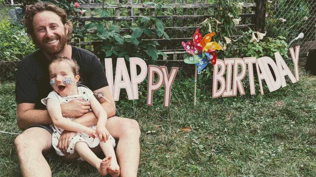 Man with beard holding young girl in lap with Happy Birthday sign behind
