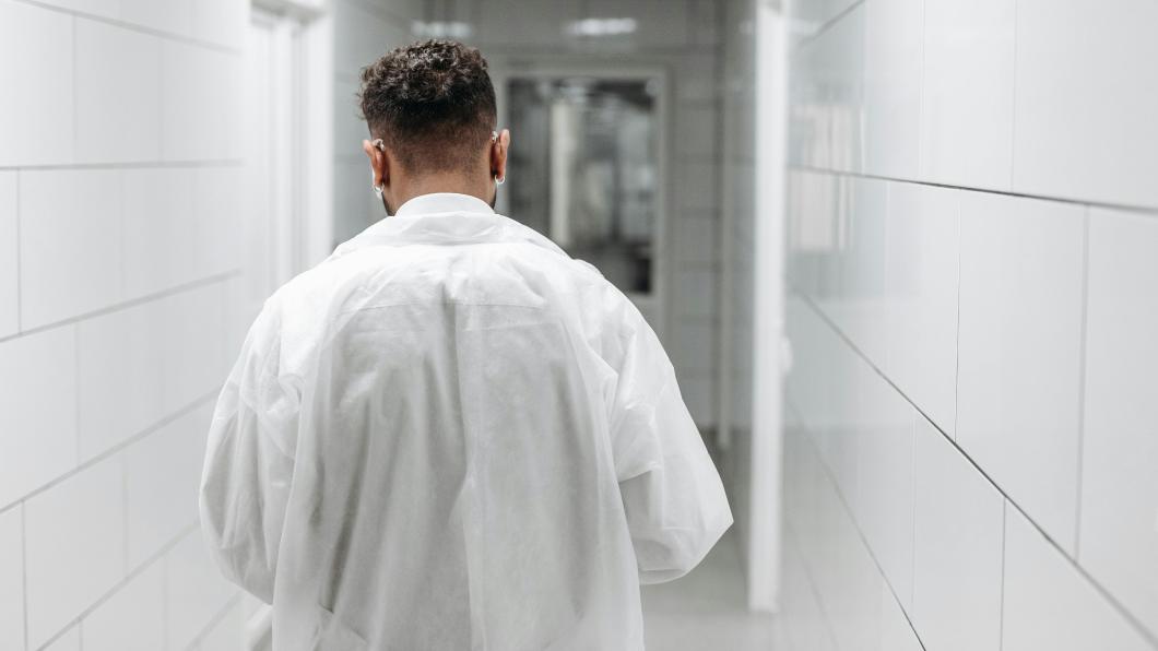 Back of man wearing white coat walking down hospital hallway.